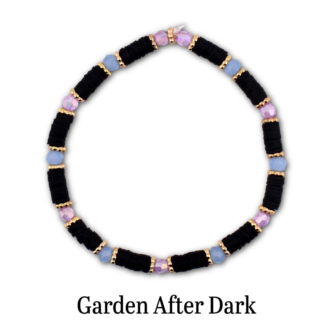 A circular beaded bracelet featuring matte black disc beads, light blue round beads, and iridescent purple faceted beads, all separated by small gold-toned metallic spacers. The text Garden After Dark is printed below the bracelet.