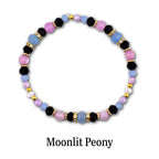 A beaded bracelet featuring a repeating pattern of black, light blue, and pink beads, interspersed with gold-toned spacers and clear crystal-encrusted rings. The text Moonlit Peony is printed below the circular jewelry piece.