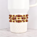 Four beaded bracelets in shades of burgundy, gold, and clear crystals are stacked on a white mug. The bracelets feature a mix of round and rectangular beads, with gold accents. The bracelets are likely for wrist wear.