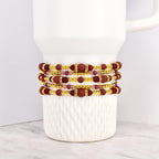 Four beaded bracelets in shades of burgundy, gold, and clear crystals are stacked on a white mug. The bracelets feature a mix of round and rectangular beads, with gold accents. The bracelets are likely for wrist wear.
