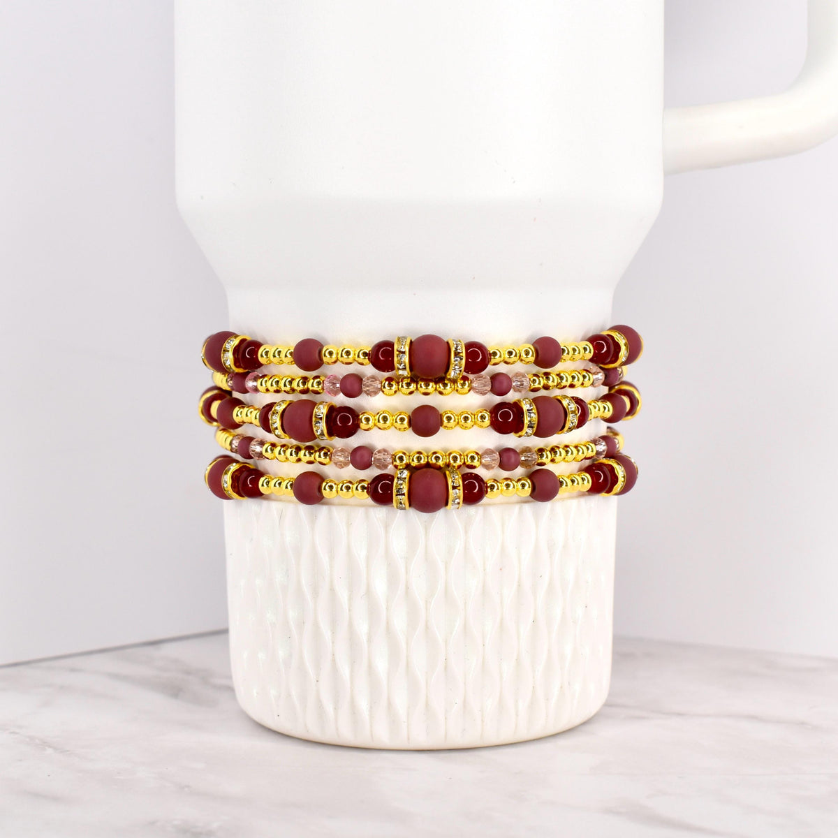 Four beaded bracelets in shades of burgundy, gold, and clear crystals are stacked on a white mug. The bracelets feature a mix of round and rectangular beads, with gold accents. The bracelets are likely for wrist wear.