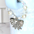 A silver bow-shaped charm with clear rhinestones hangs from a beaded bracelet of white pearls and clear, faceted beads. The charm is attached to a light blue water bottle.