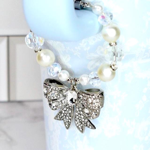 A silver bow-shaped charm with clear rhinestones hangs from a beaded bracelet of white pearls and clear, faceted beads. The charm is attached to a light blue water bottle.