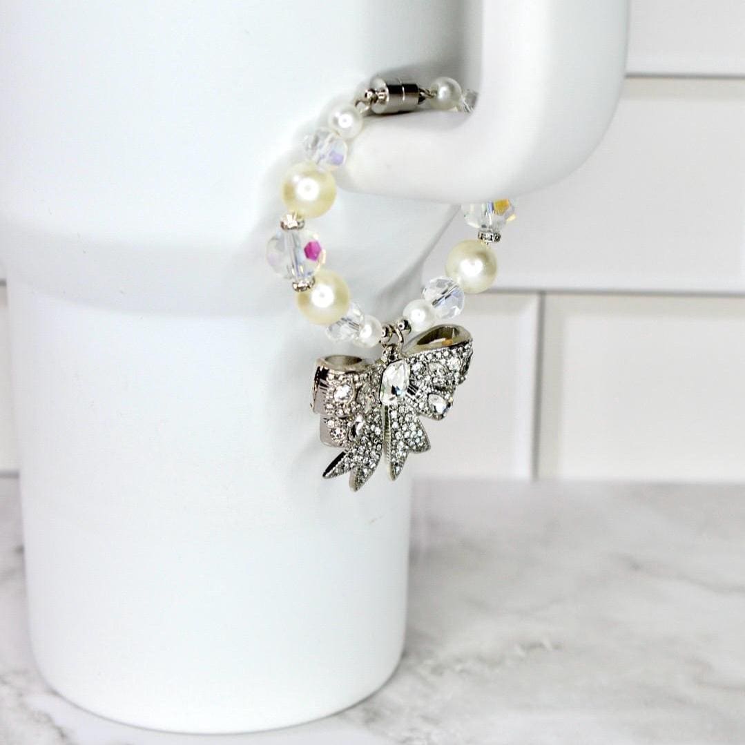 A beaded bracelet with a silver bow charm. The bracelet features a mix of white pearls, clear beads, and iridescent accents. The bow charm is encrusted with clear rhinestones. The bracelet is held by a white mug.
