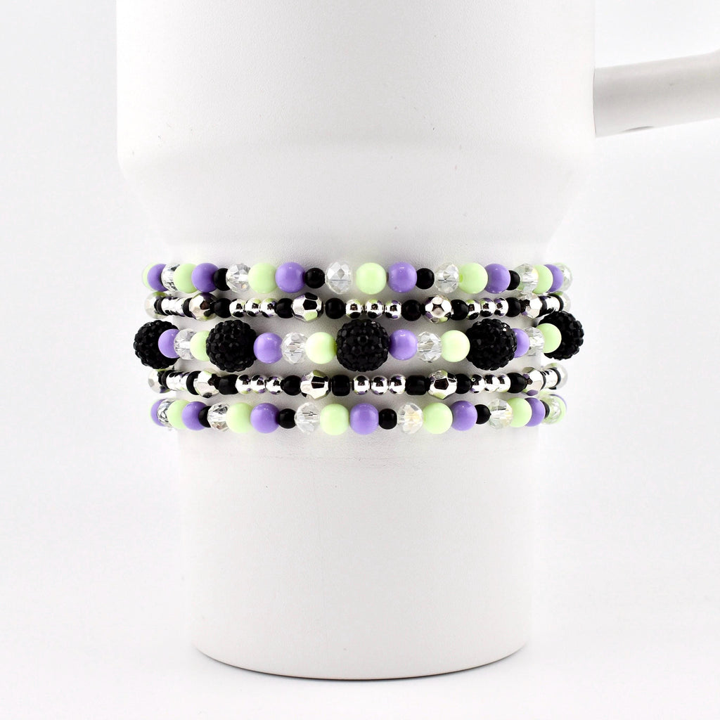 A set of beaded bracelets in shades of black, purple, green, and silver. The bracelets are made with small beads and larger, textured black beads. The bracelets are stacked on a white mug.