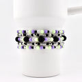 A set of beaded bracelets in shades of black, purple, green, and silver. The bracelets are made with small beads and larger, textured black beads. The bracelets are stacked on a white mug.
