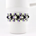 A set of beaded bracelets in shades of black, purple, green, and silver. The bracelets are made with small beads and larger, textured black beads. The bracelets are stacked on a white mug.