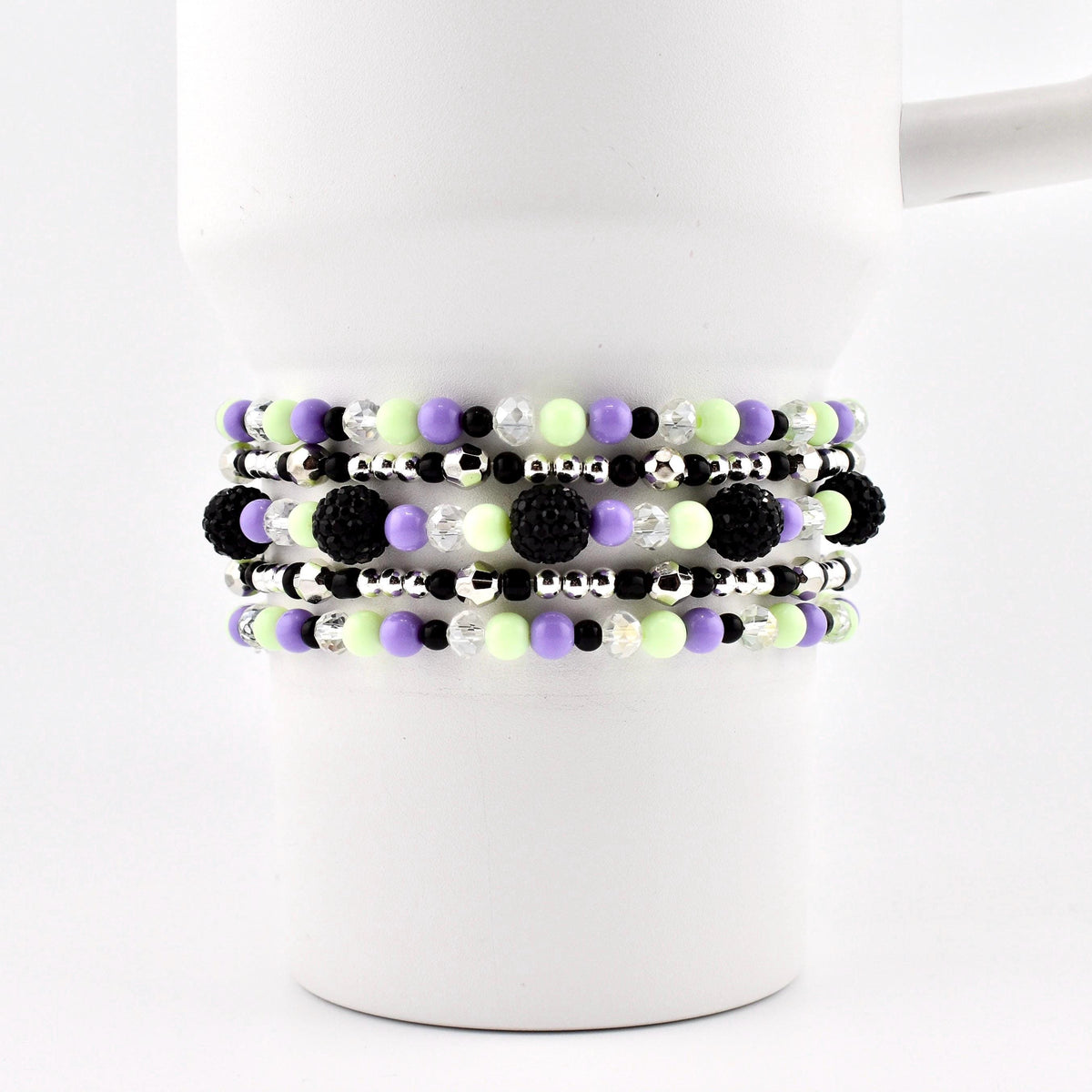 A set of beaded bracelets in shades of black, purple, green, and silver. The bracelets are made with small beads and larger, textured black beads. The bracelets are stacked on a white mug.