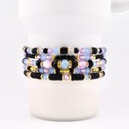 A stack of five beaded bracelets featuring a mix of black disc-shaped beads, light blue and pink translucent round beads, faceted clear crystals, and small gold-toned metallic spacers arranged in a repeating pattern around the wrist.