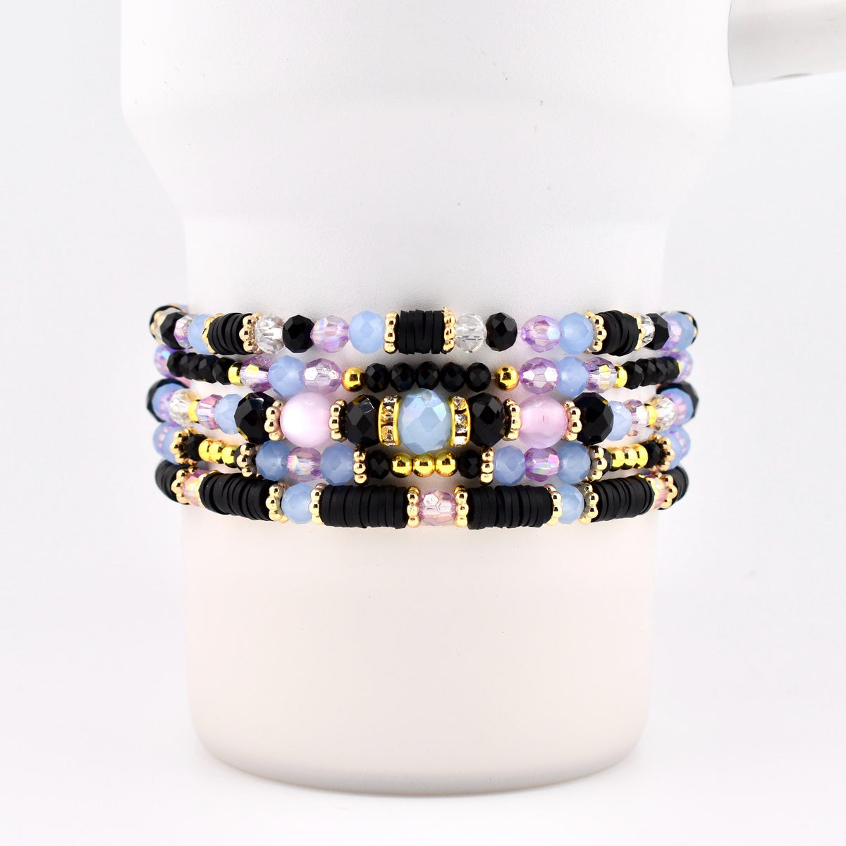 A stack of five beaded bracelets featuring a mix of black disc-shaped beads, light blue and pink translucent round beads, faceted clear crystals, and small gold-toned metallic spacers arranged in a repeating pattern around the wrist.