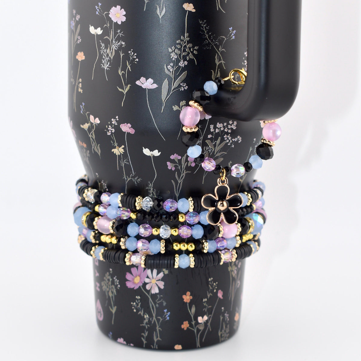 A black tumbler with a floral print featuring a stack of beaded bracelets wrapped around it. The bracelets consist of black, light blue, and pink beads with gold accents and a black flower charm hanging from the top bracelet.