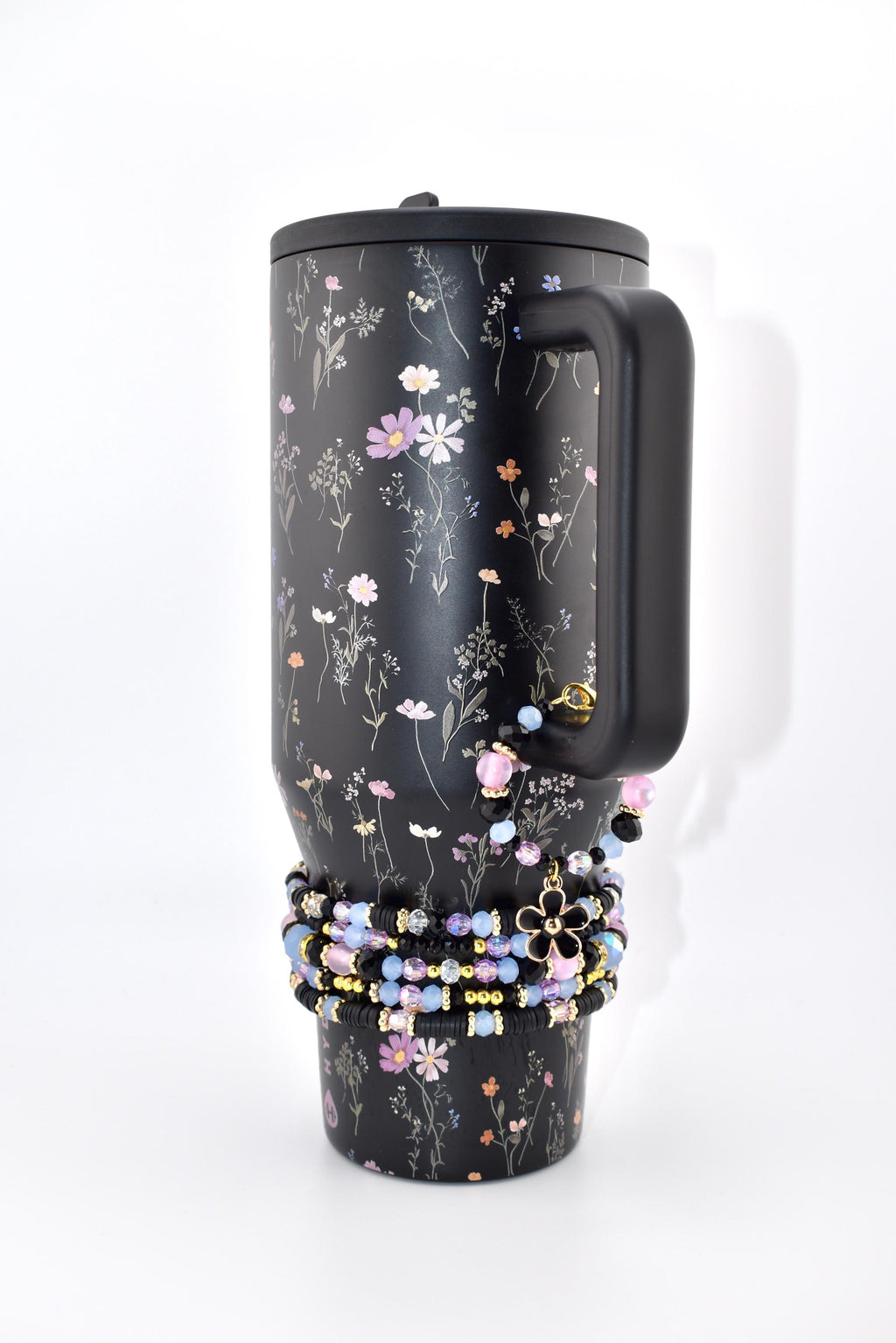 A black tumbler with a floral pattern featuring small pink, white, and orange flowers. The tumbler has a black handle and a black lid. A stack of beaded bracelets in shades of black, light blue, and pink with a black flower charm.