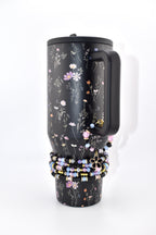 A black tumbler with a floral pattern featuring small pink, white, and orange flowers. The tumbler has a black handle and a black lid. A stack of beaded bracelets in shades of black, light blue, and pink with a black flower charm.
