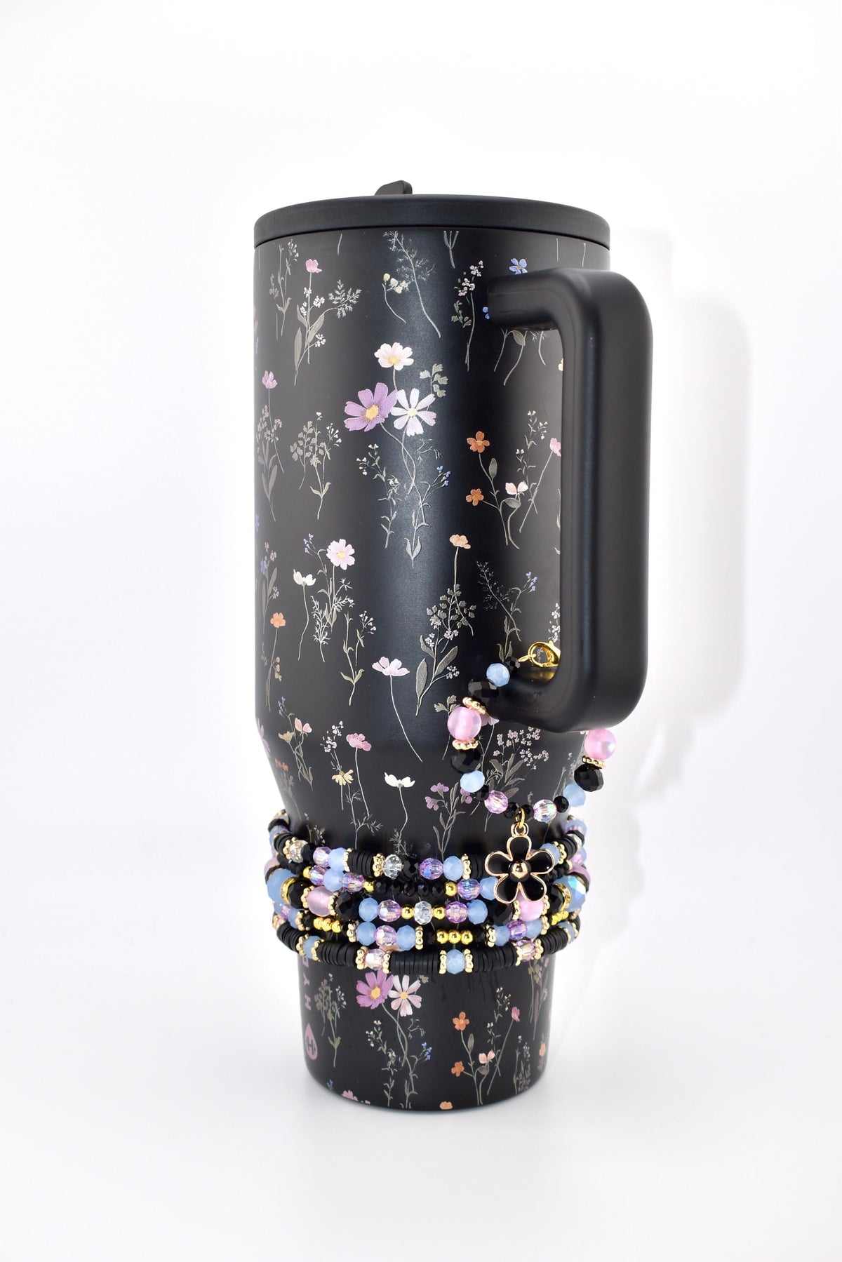 A black tumbler with a floral pattern featuring small pink, white, and orange flowers. The tumbler has a black handle and a black lid. A stack of beaded bracelets in shades of black, light blue, and pink with a black flower charm.