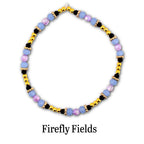 A circular beaded bracelet featuring alternating light blue and lavender faceted beads, small black beads, and metallic gold-tone spherical beads. The text Firefly Fields is printed in black below the bracelet on a white background.