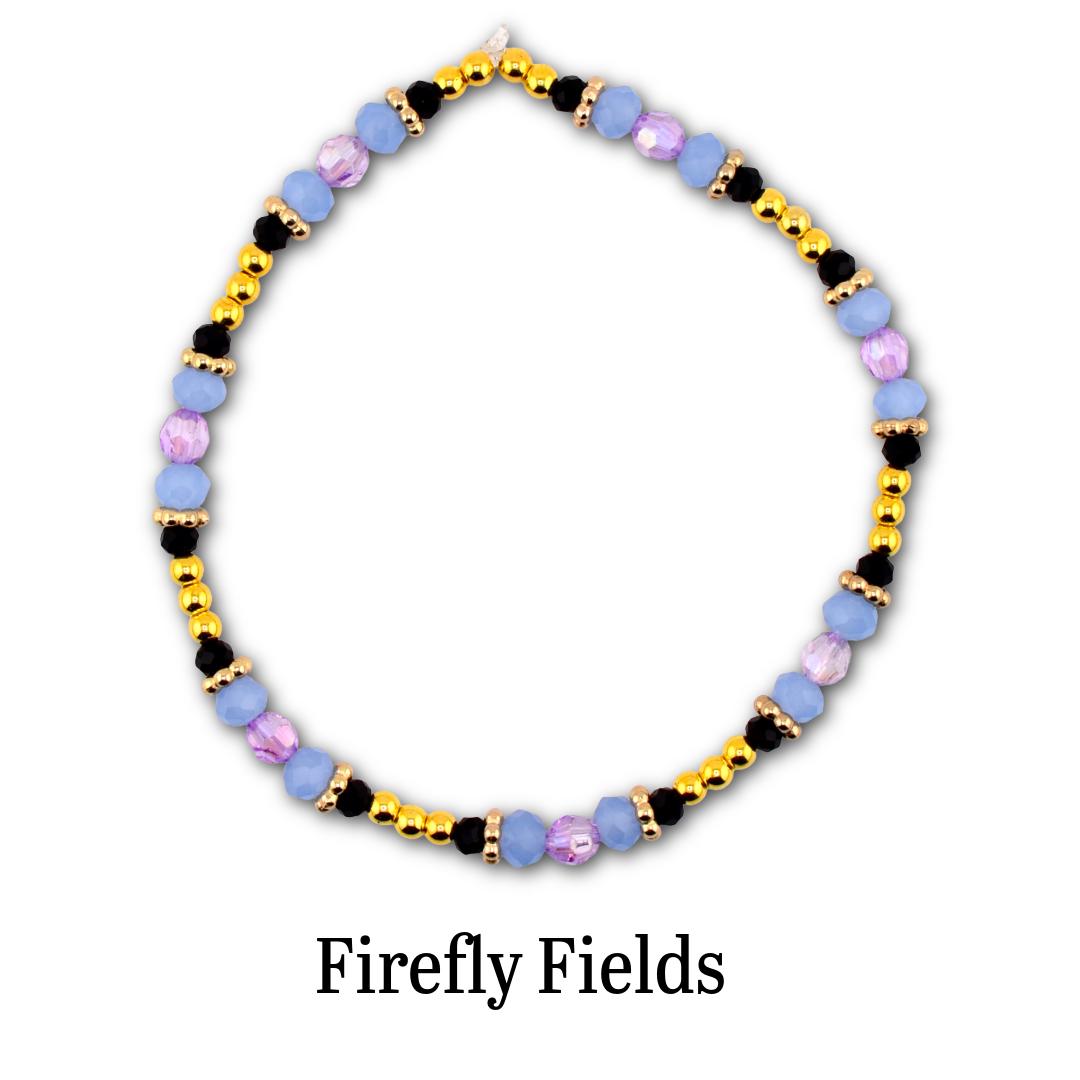 A circular beaded bracelet featuring alternating light blue and lavender faceted beads, small black beads, and metallic gold-tone spherical beads. The text Firefly Fields is printed in black below the bracelet on a white background.
