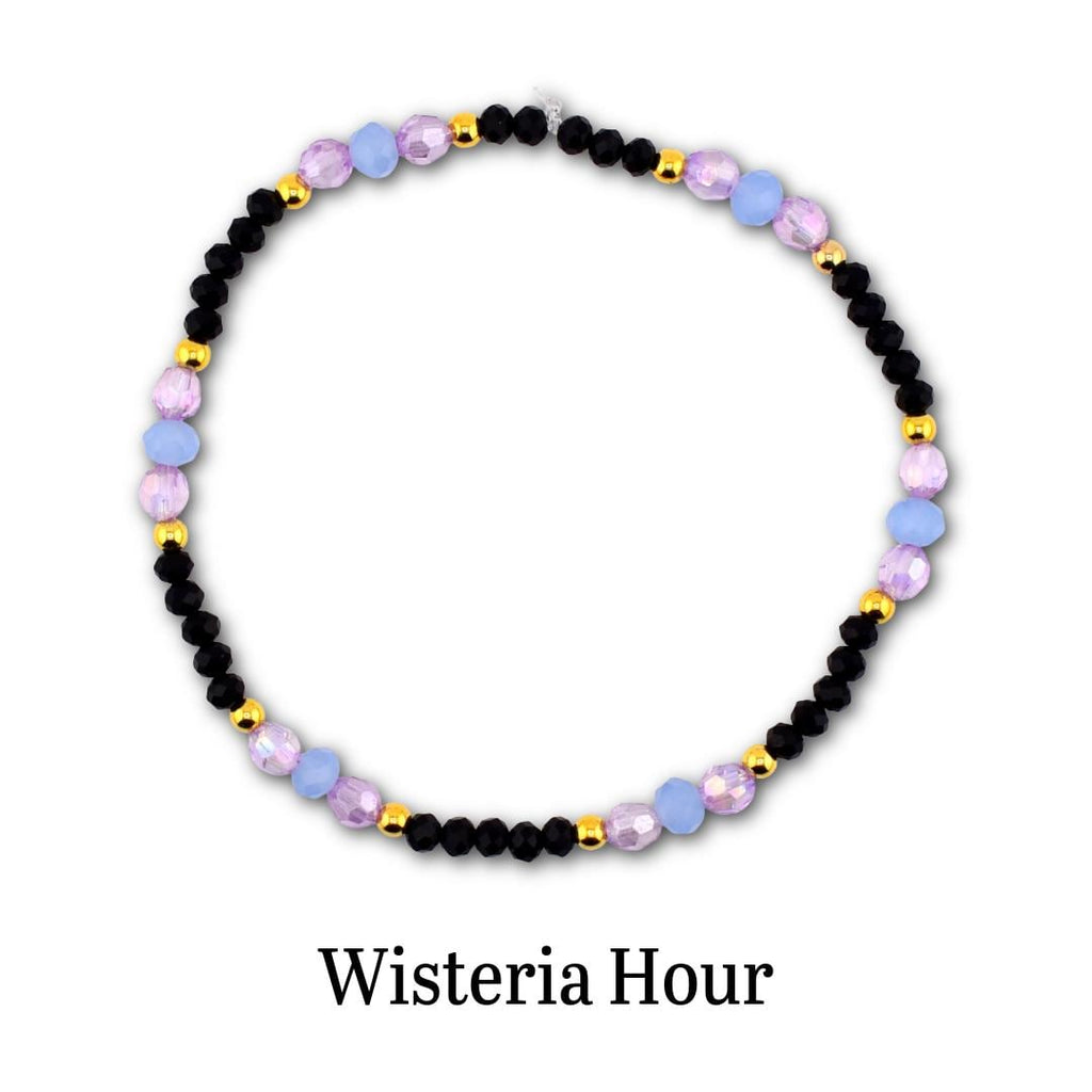 A circular beaded bracelet featuring alternating black faceted beads, light blue round beads, translucent purple beads, and small gold-toned metallic spacer beads. The text Wisteria Hour is printed in black below the bracelet.
