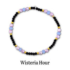 A circular beaded bracelet featuring alternating black faceted beads, light blue round beads, translucent purple beads, and small gold-toned metallic spacer beads. The text Wisteria Hour is printed in black below the bracelet.