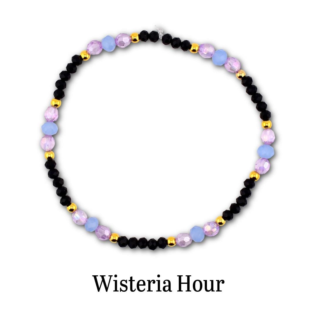 A circular beaded bracelet featuring alternating black faceted beads, light blue round beads, translucent purple beads, and small gold-toned metallic spacer beads. The text Wisteria Hour is printed in black below the bracelet.