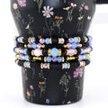 A stack of five beaded bracelets featuring a mix of black disc-shaped beads, light blue faceted crystal beads, iridescent pink round beads, and small gold-toned spacers. The bracelets are arranged in a vertical column on a black surface.
