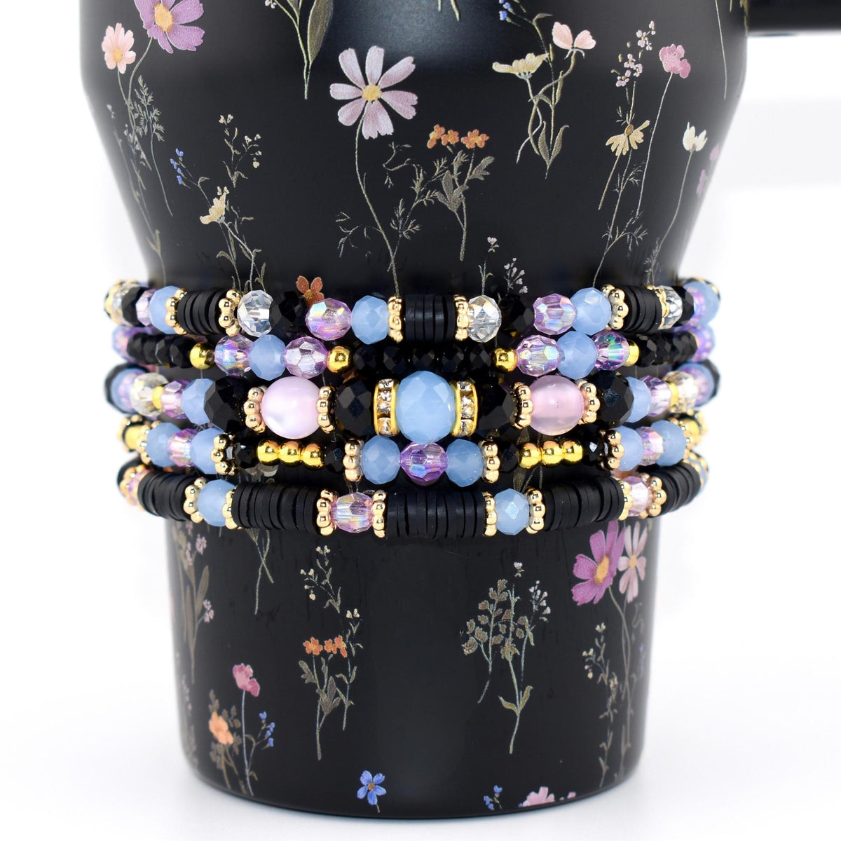 A stack of five beaded bracelets featuring a mix of black disc-shaped beads, light blue faceted crystal beads, iridescent pink round beads, and small gold-toned spacers. The bracelets are arranged in a vertical column on a black surface.