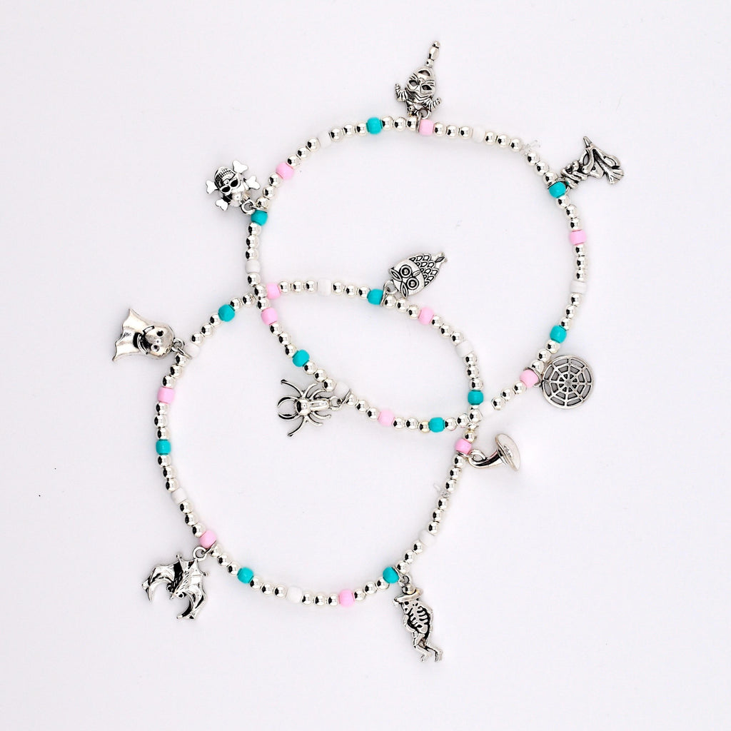 Two beaded bracelets featuring silver-toned charms including a skull, spider, bat, skeleton, and spiderweb. The bracelets are constructed with small white, pink, and turquoise beads interspersed with metallic silver spacer beads.