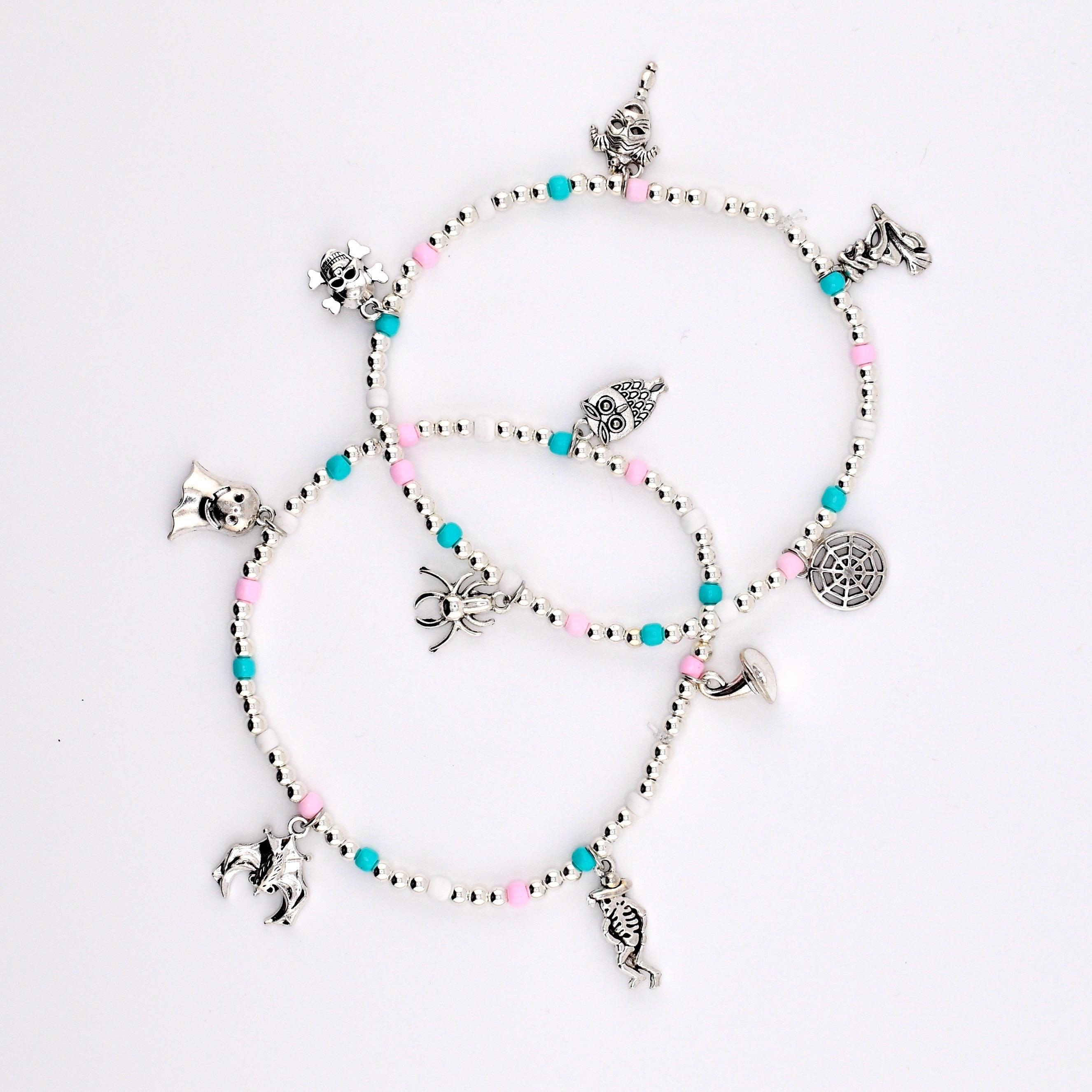 Two beaded bracelets featuring silver-toned charms including a skull, spider, bat, skeleton, and spiderweb. The bracelets are constructed with small white, pink, and turquoise beads interspersed with metallic silver spacer beads.
