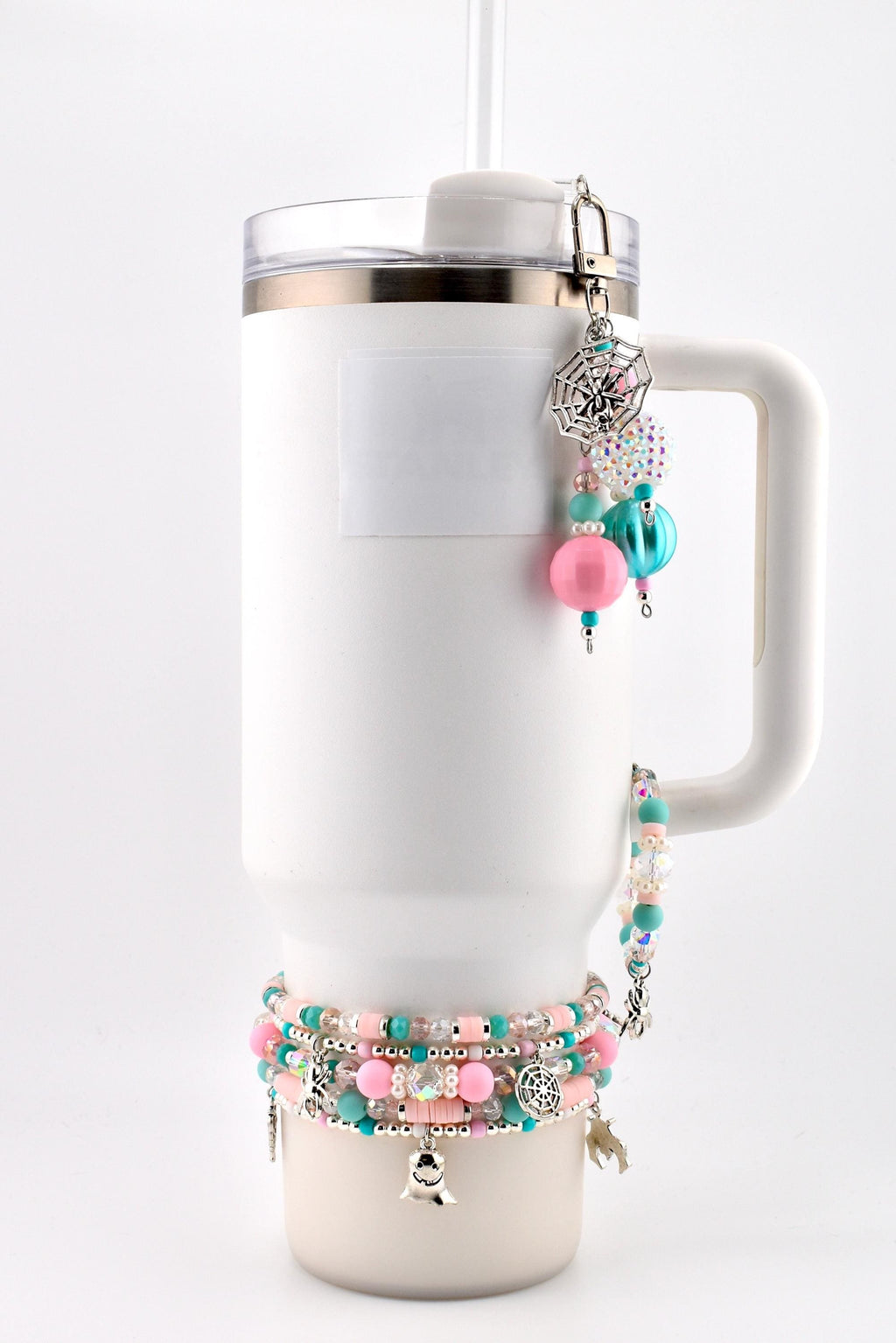 A white insulated tumbler with a handle, featuring a beaded charm attached to the lid and a stack of beaded bracelets around the base. The charms include a silver spiderweb, a ghost, and various pink, teal, and clear beads.