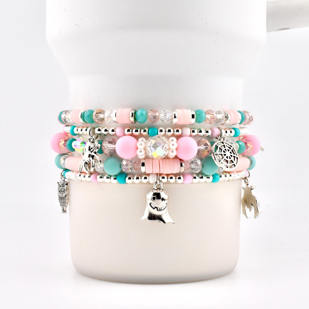 A stack of five beaded bracelets featuring pastel pink and turquoise clay discs, clear faceted beads, and round pink beads. Silver charms include a ghost, a spider, a spiderweb, an owl, and a bat, all strung on elastic cord.