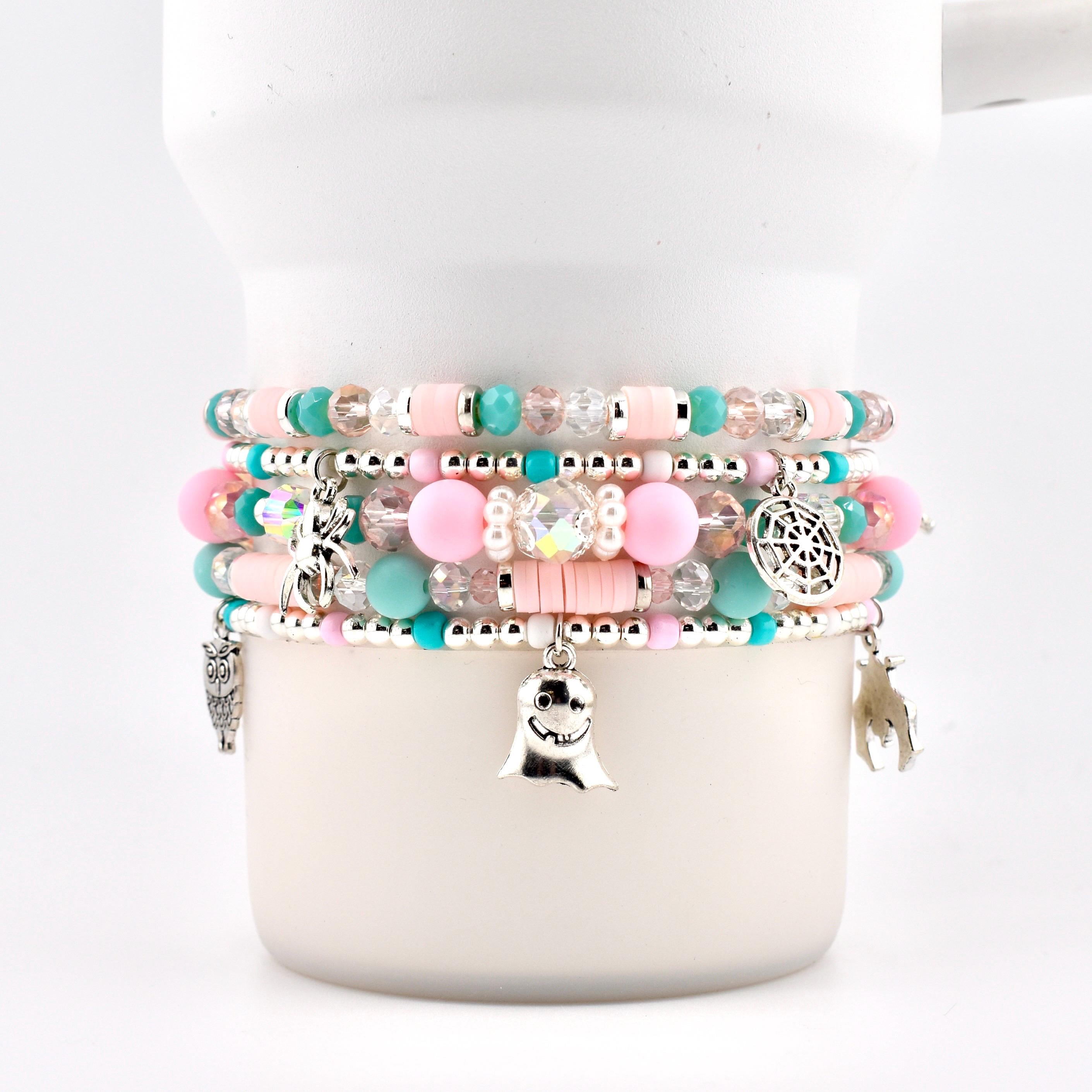 A stack of five beaded bracelets featuring pastel pink and turquoise clay discs, clear faceted beads, and round pink beads. Silver charms include a ghost, a spider, a spiderweb, an owl, and a bat, all strung on elastic cord.