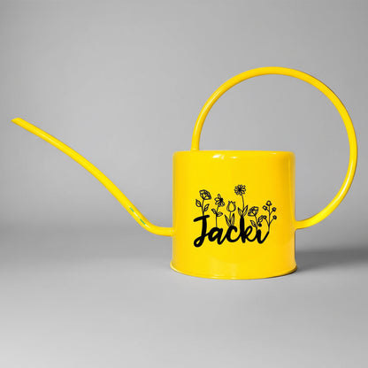 Yellow watering can with 'Jacki' and floral designs on a pink background