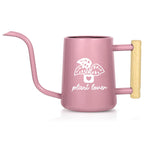 A pink watering can with a long, slender spout and a wooden handle. The can features a white graphic of a plant in a pot with the words "plant lover" below it. The watering can is designed for indoor plant care.